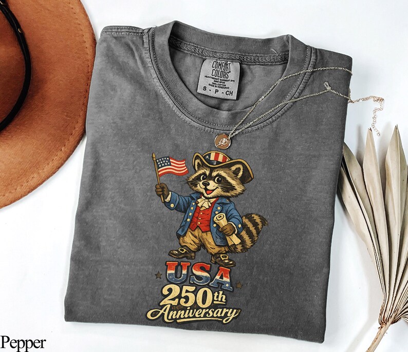 Comfort Colors 1717 t-shirt in Pepper grey featuring a vintage-style patriotic raccoon dressed as a Founding Father for the USA 250th Anniversary. The graphic shows a colonial 'Trash Panda' in a Revolutionary War uniform holding an American flag with text reading 'USA 250th Anniversary'. Perfect retro semiquincentennial gift for history buffs and Independence Day 2026 celebrations.