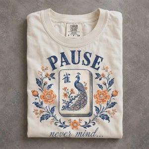 May include: A cream-colored t-shirt featuring a graphic of a mahjong tile with a peacock illustration and the Chinese character for bird. The design includes the text PAUSE and never mind, surrounded by blue and orange floral vine patterns.