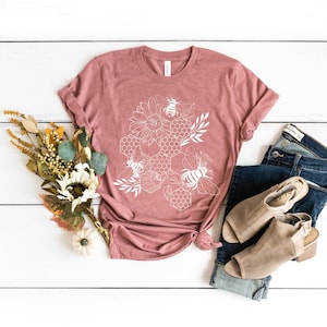May include: A mauve t-shirt with a white graphic design of a honeycomb with bees and sunflowers.