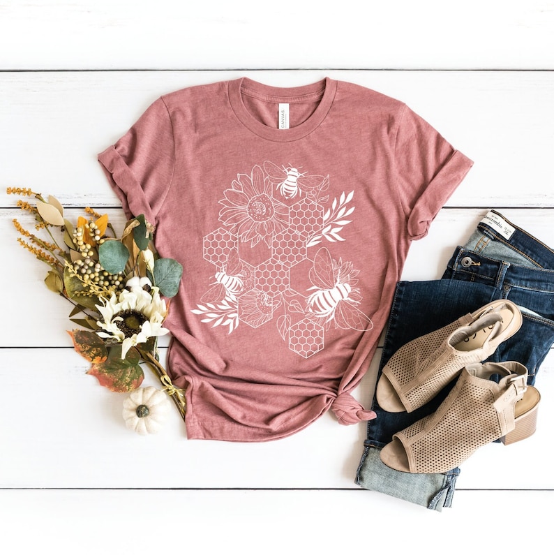 May include: A mauve t-shirt with a white graphic design of a honeycomb with bees and sunflowers.