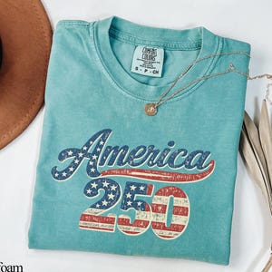 Pode incluir: Camiseta verde-&aacute;gua com a palavra "America" em estrelas e listras azuis e brancas, e o n&uacute;mero "250" em listras vermelhas e brancas. Um colar de ouro e um chap&eacute;u marrom tamb&eacute;m est&atilde;o na imagem.