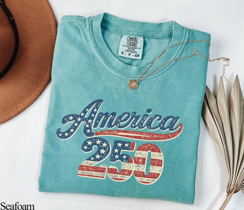Pode incluir: Camiseta verde-&aacute;gua com a palavra "America" em estrelas e listras azuis e brancas, e o n&uacute;mero "250" em listras vermelhas e brancas. Um colar de ouro e um chap&eacute;u marrom tamb&eacute;m est&atilde;o na imagem.