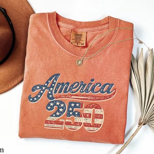 Pode incluir: Uma camiseta cor de coral com o texto "America 250" em um estilo vintage e desgastado. A palavra "America" &eacute; em azul com estrelas brancas, e o "250" apresenta um design listrado em vermelho e branco. Um colar de ouro com um pingente est&aacute; sobre a camiseta.