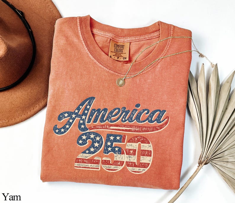 Pode incluir: Uma camiseta cor de coral com o texto "America 250" em um estilo vintage e desgastado. A palavra "America" &eacute; em azul com estrelas brancas, e o "250" apresenta um design listrado em vermelho e branco. Um colar de ouro com um pingente est&aacute; sobre a camiseta.