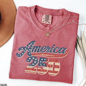Pode incluir: Camiseta carmesim com o texto "America 250" em um design patri&oacute;tico. A palavra "America" &eacute; em azul com estrelas brancas, e "250" apresenta listras vermelhas e brancas. Um colar de ouro com um pingente repousa sobre a camiseta.