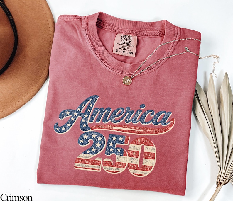 Pode incluir: Camiseta carmesim com o texto "America 250" em um design patri&oacute;tico. A palavra "America" &eacute; em azul com estrelas brancas, e "250" apresenta listras vermelhas e brancas. Um colar de ouro com um pingente repousa sobre a camiseta.