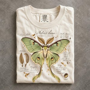 May include: A cream-colored t-shirt featuring a detailed illustration of a green Luna moth. The graphic includes scientific text labels, such as Actias luna, and notes a wingspan of 9-11 cm (3.5-4.3 in). The shirt has a crew neck and a Comfort Colors label.