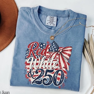 May include: A blue jean colored t-shirt with a graphic design featuring the text "Red, White & 250" in red and white, with a bow and American flag design. A gold necklace is draped across the shirt.