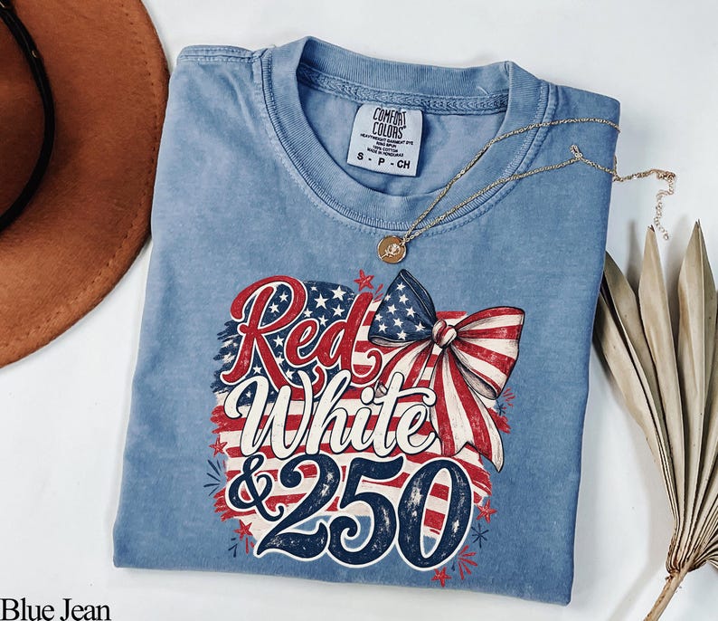 May include: A blue jean colored t-shirt with a graphic design featuring the text "Red, White & 250" in red and white, with a bow and American flag design. A gold necklace is draped across the shirt.
