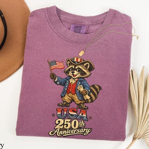 Comfort Colors 1717 Berry purple t-shirt featuring a vintage Revolutionary War raccoon in colonial attire holding an American flag. The graphic includes the text 'USA 250th Anniversary' in a retro font, making it a perfect trash panda patriot gift for the 2026 semiquincentennial celebration.