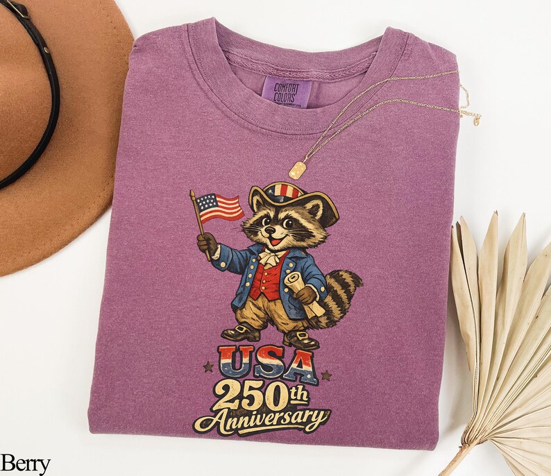 Comfort Colors 1717 Berry purple t-shirt featuring a vintage Revolutionary War raccoon in colonial attire holding an American flag. The graphic includes the text 'USA 250th Anniversary' in a retro font, making it a perfect trash panda patriot gift for the 2026 semiquincentennial celebration.