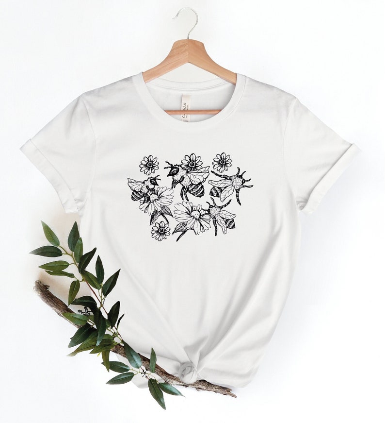 May include: White t-shirt featuring a black and white illustration of bees and flowers. The design includes several bees buzzing around a cluster of daisies. The shirt is made of a soft material and has short sleeves.