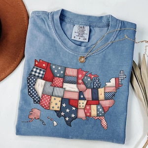 May include: A light blue Comfort Colors t-shirt featuring a patchwork design of the United States map. The map is composed of various patterns in red, white, and blue. A gold necklace with a pendant rests on the shirt.