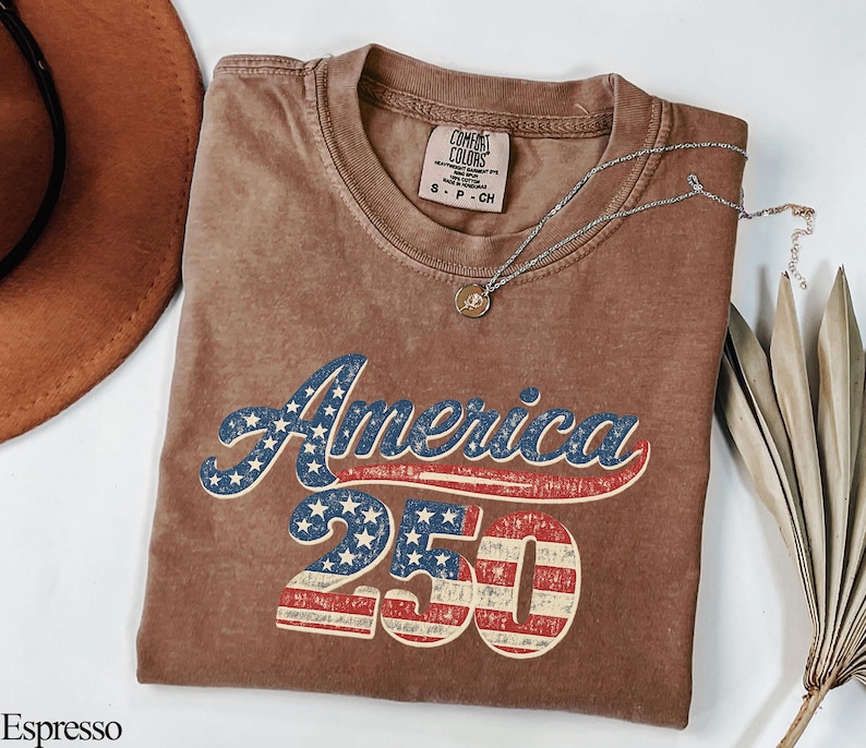Pode incluir: Camiseta cor de caf&eacute; com o texto "America 250" em um design patri&oacute;tico. A palavra "America" est&aacute; em azul com estrelas brancas, e "250" apresenta listras vermelhas e brancas. Um colar de prata com um pingente est&aacute; na camiseta.