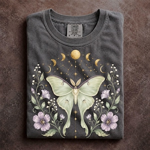 May include: A charcoal gray t-shirt featuring a central graphic of a pale green moth surrounded by purple flowers and vines. Above the moth, a series of golden moon phases and stars are displayed. The inner neck label reads Comfort Colors S P CH.