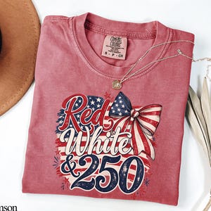 May include: Crimson-colored t-shirt with a patriotic design. The graphic features the words "Red White & 250" in a stylized font, along with a bow and stars and stripes. A gold necklace with a pendant is draped on the shirt.