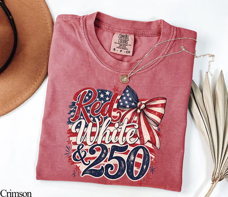 May include: Crimson-colored t-shirt with a patriotic design. The graphic features the words "Red White & 250" in a stylized font, along with a bow and stars and stripes. A gold necklace with a pendant is draped on the shirt.