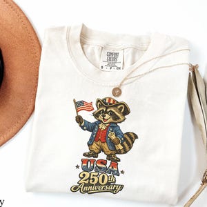 Comfort Colors 1717 Ivory t-shirt featuring a colonial founding father raccoon character holding an American flag with text reading 'USA 250th Anniversary'. This vintage-style patriotic tee is perfect for the 2026 Semiquincentennial celebration, making it a unique gift for history buffs and lovers of funny trash panda apparel.