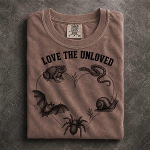 May include: A brown crew neck t-shirt featuring a black graphic print of a toad, snake, bat, snail, and spider arranged in a heart shape. The text LOVE THE UNLOVED is printed in black above the heart. The shirt has a Comfort Colors brand label.