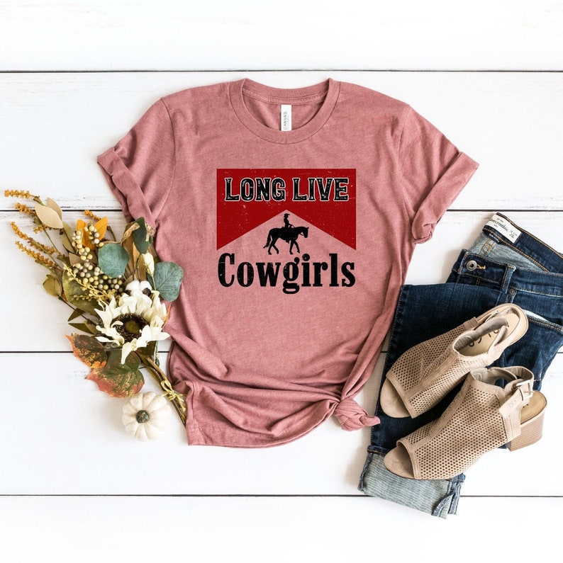 T-shirt in a heather mauve color featuring a 'Long Live Cowgirls' Marlboro parody design with a horse rider silhouette in red and black. This retro western graphic tee is styled in a flat lay with jeans and booties, perfect for a country concert outfit or Nashville bachelorette trip.