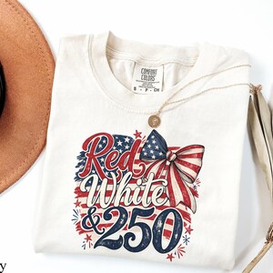 May include: Ivory-colored t-shirt with a patriotic design. The graphic features the words "Red White & 250" in red and blue, with a bow and stars. A gold necklace and a brown hat are also visible.