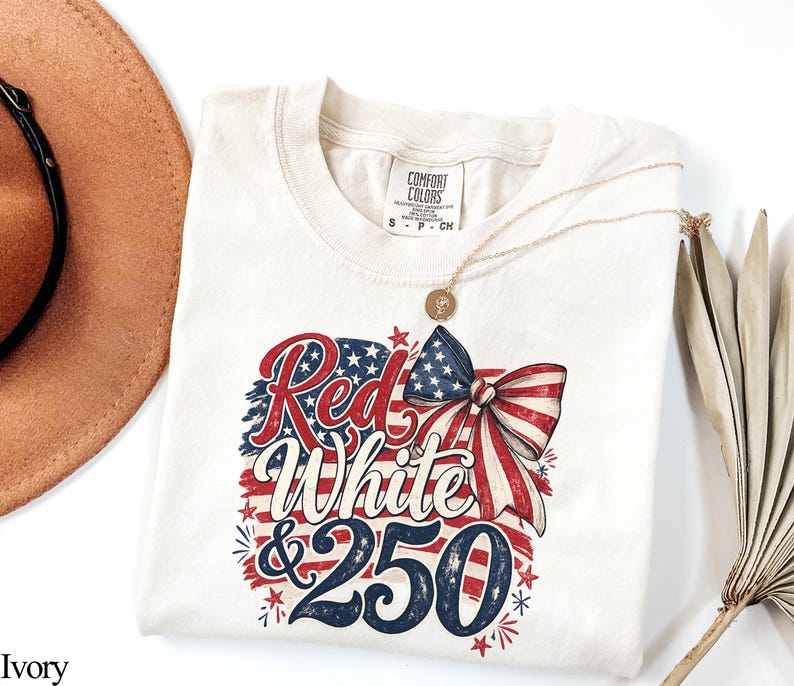 May include: Ivory-colored t-shirt with a patriotic design. The graphic features the words "Red White & 250" in red and blue, with a bow and stars. A gold necklace and a brown hat are also visible.
