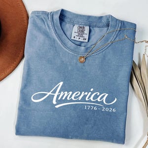 May include: A blue t-shirt with the word "America" in white script and the numbers "1776-2026" printed below. A gold necklace with a pendant is draped across the shirt. A brown hat and dried plant are in the background.