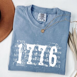 May include: A blue t-shirt with the number "1776" in large white letters. The shirt has a crew neck and short sleeves. A gold necklace with a pendant is draped on the shirt. The shirt is folded on a white surface.