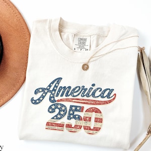 Pode incluir: Camiseta de cor marfim com o texto "America 250" em um design vintage e patri&oacute;tico. A palavra "America" est&aacute; em azul com estrelas brancas, e "250" apresenta listras e estrelas vermelhas e brancas. Um colar dourado e um chap&eacute;u marrom tamb&eacute;m s&atilde;o vis&iacute;veis.