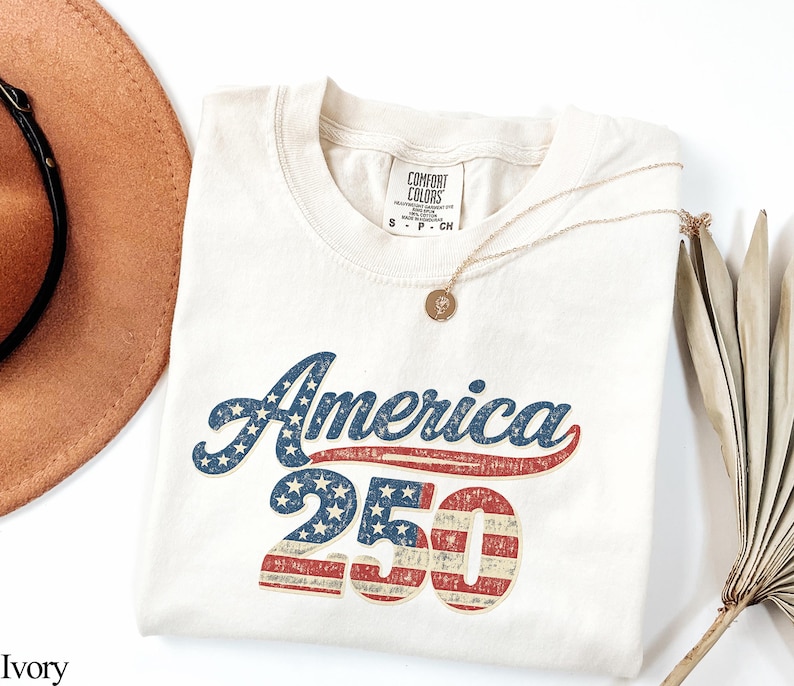 Pode incluir: Camiseta de cor marfim com o texto "America 250" em um design vintage e patri&oacute;tico. A palavra "America" est&aacute; em azul com estrelas brancas, e "250" apresenta listras e estrelas vermelhas e brancas. Um colar dourado e um chap&eacute;u marrom tamb&eacute;m s&atilde;o vis&iacute;veis.