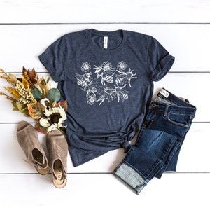 May include: A navy blue t-shirt with a white floral and bee design. The shirt is paired with blue jeans and beige open-toe shoes. A bouquet of fall-colored flowers and foliage is arranged on a white wooden surface.