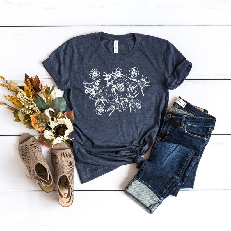 May include: A navy blue t-shirt with a white floral and bee design. The shirt is paired with blue jeans and beige open-toe shoes. A bouquet of fall-colored flowers and foliage is arranged on a white wooden surface.