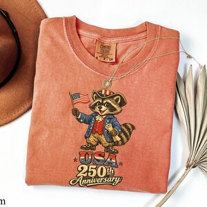 Comfort Colors 1717 garment-dyed shirt in Yam featuring a vintage-style raccoon dressed in a colonial Revolutionary War uniform. The design includes a 'Trash Panda Patriot' holding an American flag with 'USA 250th Anniversary' text, making it a perfect retro patriotic gift for the 2026 Semiquincentennial celebration.