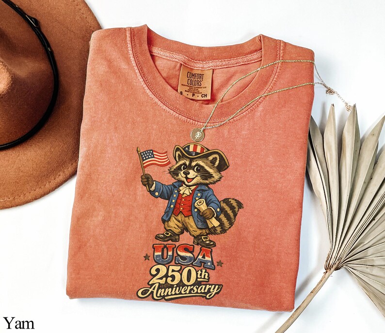 Comfort Colors 1717 garment-dyed shirt in Yam featuring a vintage-style raccoon dressed in a colonial Revolutionary War uniform. The design includes a 'Trash Panda Patriot' holding an American flag with 'USA 250th Anniversary' text, making it a perfect retro patriotic gift for the 2026 Semiquincentennial celebration.