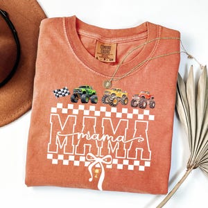 May include: A coral-colored t-shirt with a graphic design featuring monster trucks and the word "MAMA" in a checkered pattern. The shirt has a crew neck and short sleeves. A gold necklace is draped across the shirt.