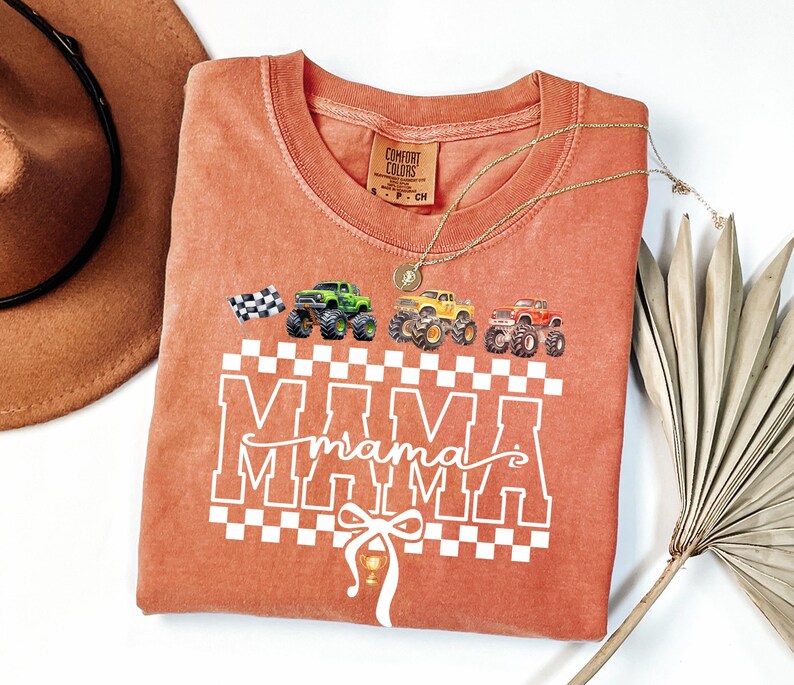 May include: A coral-colored t-shirt with a graphic design featuring monster trucks and the word "MAMA" in a checkered pattern. The shirt has a crew neck and short sleeves. A gold necklace is draped across the shirt.