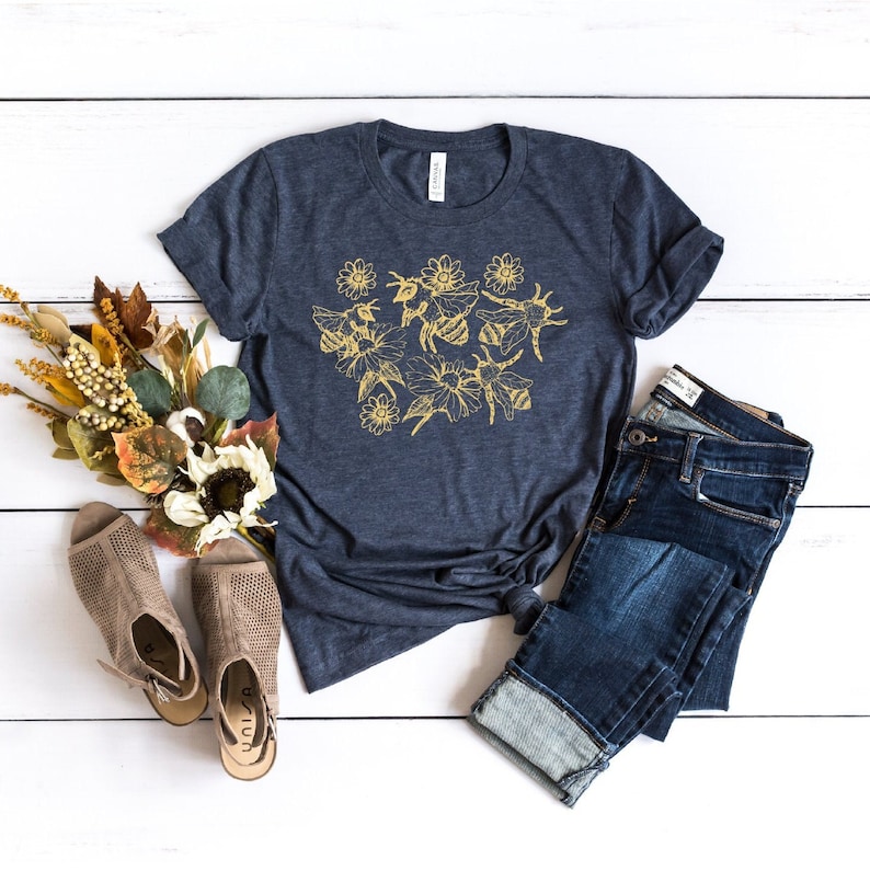 May include: A navy blue t-shirt with a gold floral and bee design. The shirt is paired with blue jeans and tan open-toe shoes. A bouquet of fall-colored flowers and foliage is arranged to the left of the shirt.