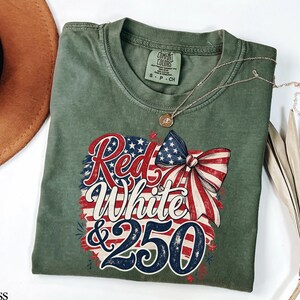 May include: Moss green t-shirt with a patriotic design. The graphic features the words "Red White & 250" in red, white, and blue, along with a bow and American flag elements. A gold necklace is draped over the shirt.