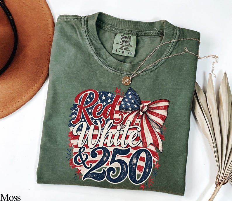 May include: Moss green t-shirt with a patriotic design. The graphic features the words "Red White & 250" in red, white, and blue, along with a bow and American flag elements. A gold necklace is draped over the shirt.