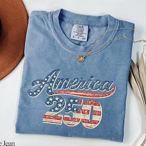 Pode incluir: Camiseta azul jeans com a inscri&ccedil;&atilde;o "America 250" em um design patri&oacute;tico. A camiseta tem gola redonda e mangas curtas. Um colar de ouro com um pingente est&aacute; drapeado sobre a camiseta. Um chap&eacute;u marrom e uma planta seca est&atilde;o no fundo.