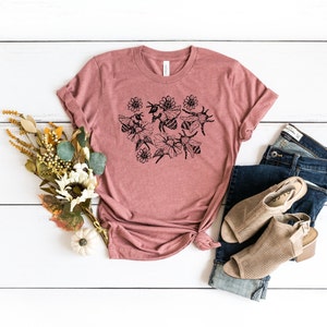 May include: A dusty rose t-shirt featuring a black floral and bee design. The shirt is styled with blue jeans and tan open-toe booties. A bouquet of fall-colored flowers and a small white pumpkin are also in the image.