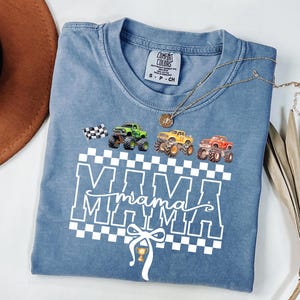 May include: A blue Comfort Colors t-shirt featuring a graphic of monster trucks in green, yellow, and red, with a checkered flag and the word "MAMA" in white. A gold necklace and a brown hat are also visible.