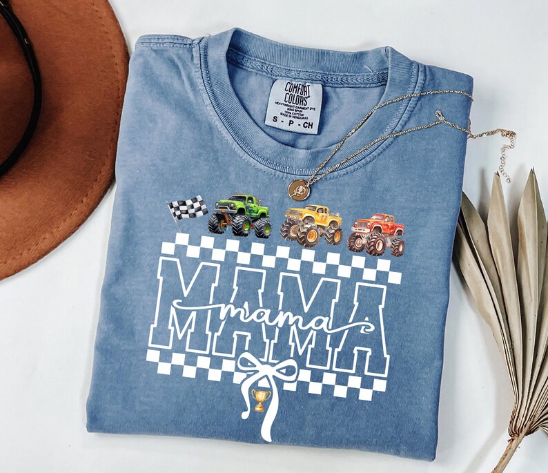 May include: A blue Comfort Colors t-shirt featuring a graphic of monster trucks in green, yellow, and red, with a checkered flag and the word "MAMA" in white. A gold necklace and a brown hat are also visible.