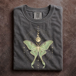 May include: A charcoal gray t-shirt featuring a graphic of a pale green luna moth centered below a vertical sequence of moon phases and a full moon. The design includes thin gold circular lines and small starburst accents. The neck label reads Comfort Colors.