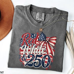 May include: A gray t-shirt with the words "Red, White & 250" in a patriotic design. The design includes a bow and the American flag colors of red, white, and blue. The shirt is made of a soft material and has a round neckline.