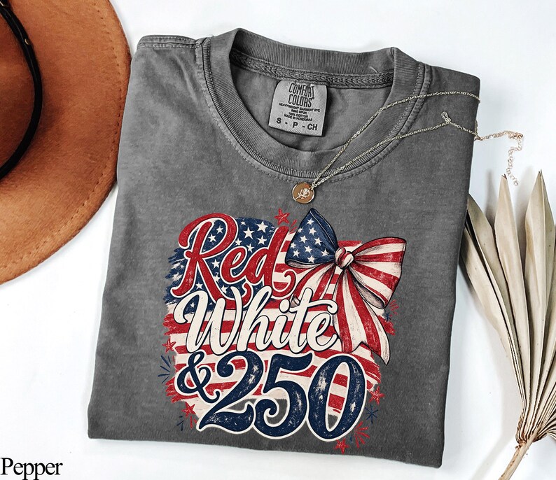 May include: A gray t-shirt with the words "Red, White & 250" in a patriotic design. The design includes a bow and the American flag colors of red, white, and blue. The shirt is made of a soft material and has a round neckline.
