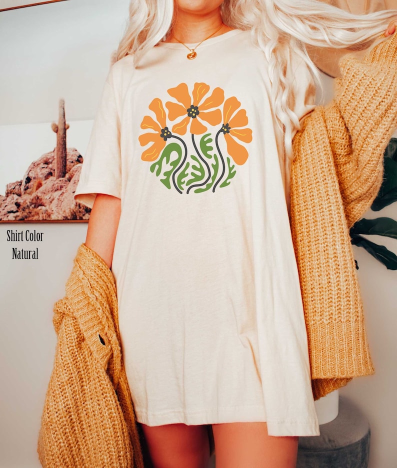 Op de afbeelding: Een wit oversized T-shirt met een afbeelding van drie oranje bloemen met groene stelen en bladeren. Het shirt wordt gedragen door een persoon met lang blond haar.