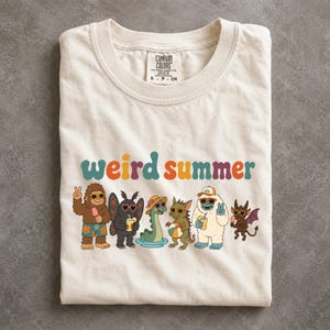 May include: A cream-colored t-shirt featuring the text "weird summer" in colorful letters above a graphic of six cartoon creatures wearing sunglasses and summer accessories, including a yeti, a moth, a sea monster, and a small dragon.