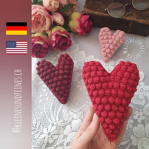May include: Crocheted hearts in varying shades of red and pink, featuring a textured, bobbled design. The hearts are arranged on a white lace surface, with pink roses and vintage spectacles in the background. The image includes the text @KLEINESUNDFEINES.CH.