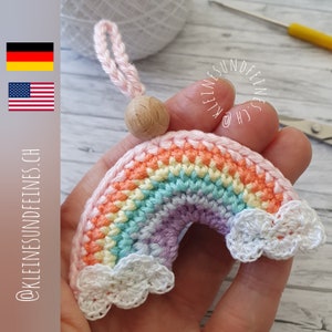 May include: A crocheted rainbow with clouds, in shades of pink, orange, yellow, green, blue, and purple, with a wooden bead at the top. The rainbow is attached to a pink string.
